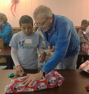 RSVP volunteer mentor, David Hinds, and his mentee, CJ, try to wrap a gift box with only one hand each during game time at a holiday party held in December for the Outagamie County Mentoring Program. 